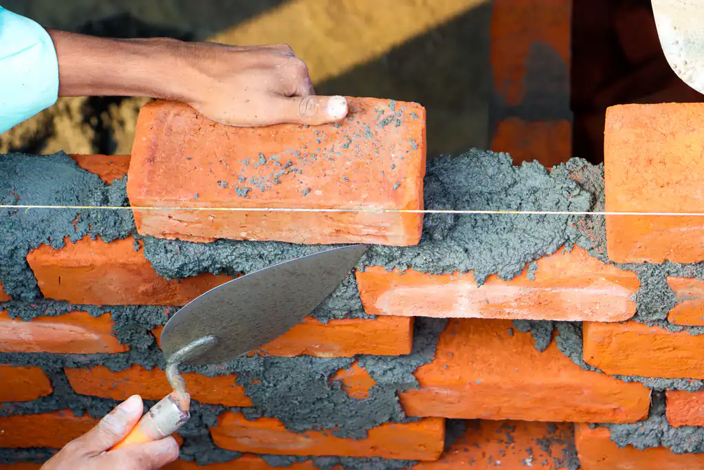 Close-up of a bricklayer laying a red brick to a line, trowel in hand
