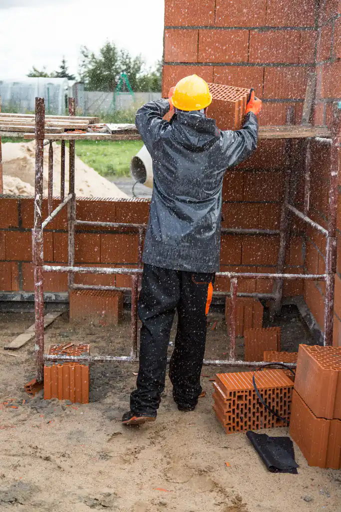 Builder in a hard hat laying clay blocks from a scaffold in the rain
